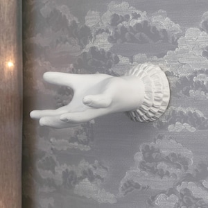 May include: A white wall-mounted hook shaped like a human hand with an open palm and fingers extended. The base of the hand features a decorative ruffled cuff detail, providing a functional and sculptural element for hanging items on a wall.