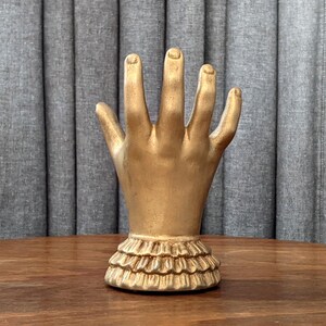 K&ouml;nnte beinhalten: Eine goldfarbene Handskulptur mit ausgestreckten Fingern. Die Hand ist auf einem ger&uuml;schten Sockel montiert. Die Skulptur wird auf einer Holzoberfl&auml;che vor einem grauen Vorhanghintergrund ausgestellt.