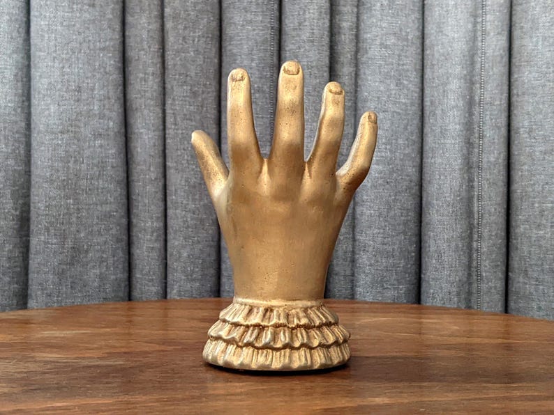 K&ouml;nnte beinhalten: Eine goldfarbene Handskulptur mit ausgestreckten Fingern. Die Hand ist auf einem ger&uuml;schten Sockel montiert. Die Skulptur wird auf einer Holzoberfl&auml;che vor einem grauen Vorhanghintergrund ausgestellt.