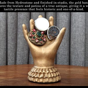 K&ouml;nnte beinhalten: Eine goldfarbene Handskulptur, die drei dekorative Ringe h&auml;lt. Die Ringe umfassen einen Ring mit wei&szlig;em Stein, eine Schmuckbrosche und einen Fossilring. Die Skulptur hat einen ger&uuml;schten Sockel. Der Text im Bild lautet: Hergestellt aus Hydrostone und im Studio fertiggestellt, f&auml;ngt die goldene Hand die Textur und Patina einer echten Antiquit&auml;t ein und verleiht ihr eine warme, taktile Pr&auml;senz, die historisch und einzigartig wirkt.