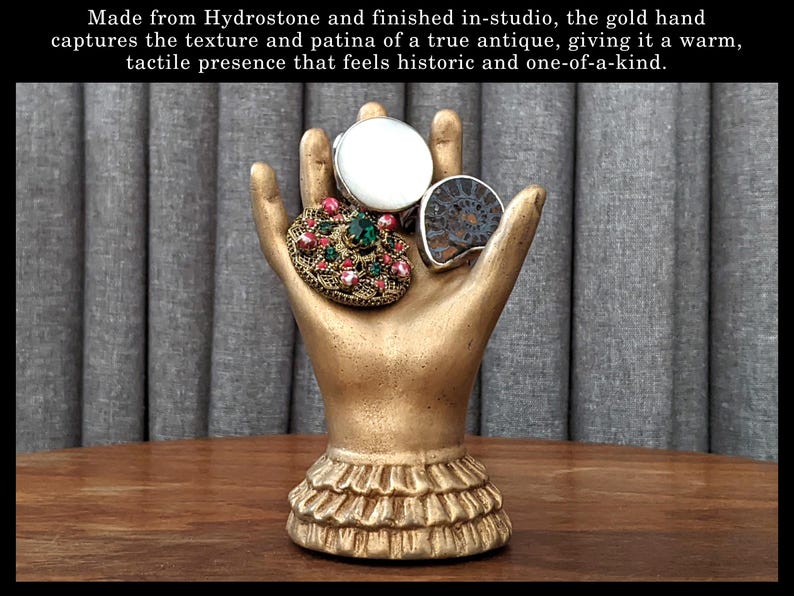 K&ouml;nnte beinhalten: Eine goldfarbene Handskulptur, die drei dekorative Ringe h&auml;lt. Die Ringe umfassen einen Ring mit wei&szlig;em Stein, eine Schmuckbrosche und einen Fossilring. Die Skulptur hat einen ger&uuml;schten Sockel. Der Text im Bild lautet: Hergestellt aus Hydrostone und im Studio fertiggestellt, f&auml;ngt die goldene Hand die Textur und Patina einer echten Antiquit&auml;t ein und verleiht ihr eine warme, taktile Pr&auml;senz, die historisch und einzigartig wirkt.