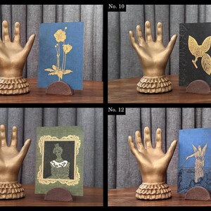K&ouml;nnte beinhalten: Eine Sammlung von vier dekorativen Hand-Display-St&auml;ndern. Jede goldfarbene Hand hat einen ger&uuml;schten Sockel und h&auml;lt eine Karte. Die Karten zeigen verschiedene Designs, darunter Blumen- und Figurenillustrationen, in Gold und anderen Farben.