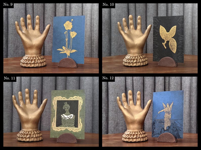K&ouml;nnte beinhalten: Eine Sammlung von vier dekorativen Hand-Display-St&auml;ndern. Jede goldfarbene Hand hat einen ger&uuml;schten Sockel und h&auml;lt eine Karte. Die Karten zeigen verschiedene Designs, darunter Blumen- und Figurenillustrationen, in Gold und anderen Farben.