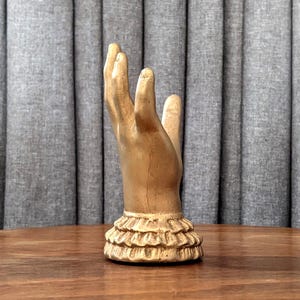 K&ouml;nnte beinhalten: Eine goldfarbene Handskulptur mit ger&uuml;schtem Sockel. Die Hand ist mit nach oben ausgestreckten Fingern dargestellt. Die Skulptur steht auf einer Holzoberfl&auml;che vor einem grauen Vorhang.