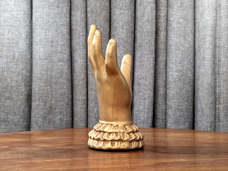 K&ouml;nnte beinhalten: Eine goldfarbene Handskulptur mit ger&uuml;schtem Sockel. Die Hand ist mit nach oben ausgestreckten Fingern dargestellt. Die Skulptur steht auf einer Holzoberfl&auml;che vor einem grauen Vorhang.
