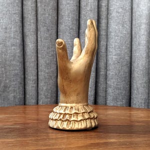 K&ouml;nnte beinhalten: Eine goldfarbene Handskulptur mit erhobenem Zeige- und Mittelfinger. Der Sockel der Skulptur hat ein R&uuml;schendesign. Die Skulptur steht auf einer Holzoberfl&auml;che mit einem grauen Vorhang im Hintergrund.