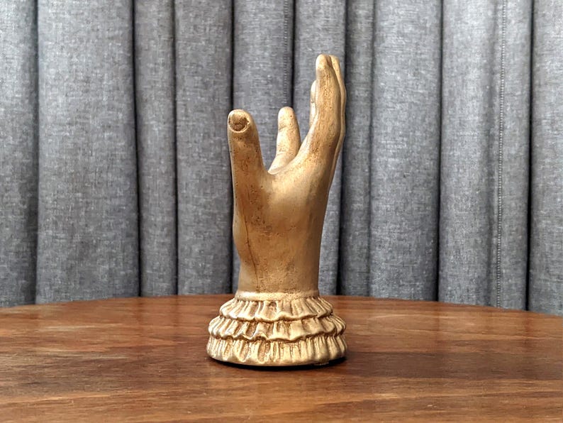 K&ouml;nnte beinhalten: Eine goldfarbene Handskulptur mit erhobenem Zeige- und Mittelfinger. Der Sockel der Skulptur hat ein R&uuml;schendesign. Die Skulptur steht auf einer Holzoberfl&auml;che mit einem grauen Vorhang im Hintergrund.
