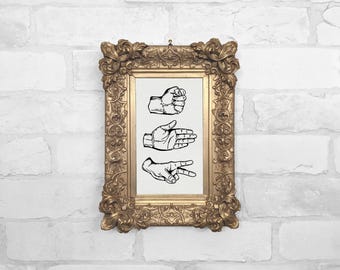 May include: A framed print showing the rock, paper, scissors hand gestures. The black and white illustrations are set against a cream background, within an ornate, gold-coloured frame. The frame hangs on a white brick wall.
