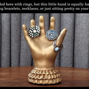 K&ouml;nnte beinhalten: Ein goldfarbener, handf&ouml;rmiger Schmuckhalter mit drei Ringen. Die Ringe sind mit wei&szlig;en, blauen und t&uuml;rkisfarbenen Steinen verziert. Der Sockel des Halters hat ein R&uuml;schendesign. Der Text auf dem Bild lautet: "Hier mit Ringen gestylt, aber diese kleine Hand ist genauso gl&uuml;cklich, Armb&auml;nder, Halsketten zu halten oder einfach nur h&uuml;bsch auf Ihrem Regal zu sitzen."