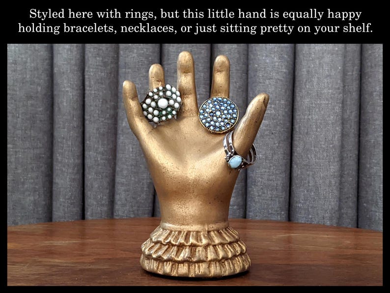 K&ouml;nnte beinhalten: Ein goldfarbener, handf&ouml;rmiger Schmuckhalter mit drei Ringen. Die Ringe sind mit wei&szlig;en, blauen und t&uuml;rkisfarbenen Steinen verziert. Der Sockel des Halters hat ein R&uuml;schendesign. Der Text auf dem Bild lautet: "Hier mit Ringen gestylt, aber diese kleine Hand ist genauso gl&uuml;cklich, Armb&auml;nder, Halsketten zu halten oder einfach nur h&uuml;bsch auf Ihrem Regal zu sitzen."