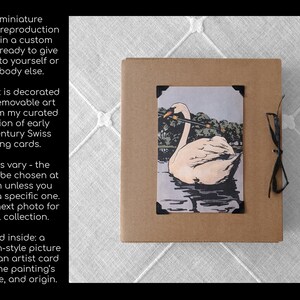 Op de afbeelding: Miniatuur schilderij reproductie in een op maat gemaakte geschenkdoos. De doos is bruin met een zwart lint. De kunstkaart toont een zwaan op water, met een zwart kader. Tekst aan de linkerkant beschrijft het product.