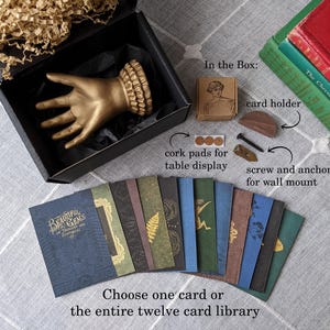 K&ouml;nnte beinhalten: Eine goldfarbene Handskulptur in einer schwarzen Box, mit einer Auswahl von zw&ouml;lf dekorativen Karten. Die Karten haben verschiedene Designs und Farben. Zus&auml;tzliche Artikel sind ein Kartenhalter, Korkuntersetzer und eine Schraube zur Wandmontage.