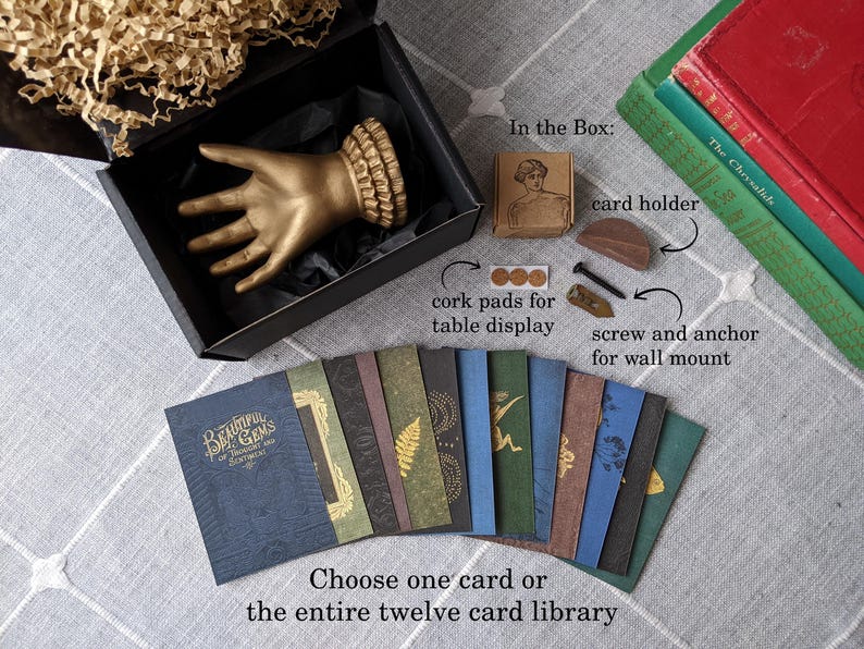 K&ouml;nnte beinhalten: Eine goldfarbene Handskulptur in einer schwarzen Box, mit einer Auswahl von zw&ouml;lf dekorativen Karten. Die Karten haben verschiedene Designs und Farben. Zus&auml;tzliche Artikel sind ein Kartenhalter, Korkuntersetzer und eine Schraube zur Wandmontage.