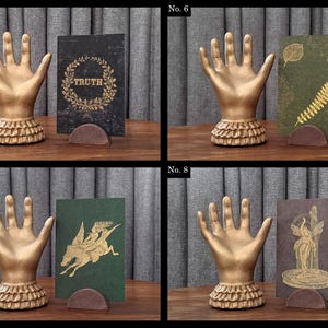 K&ouml;nnte beinhalten: Vier Bilder zeigen dekorative goldene Handskulpturen mit ger&uuml;schtem Sockel. Jede Hand h&auml;lt eine Karte in einem braunen Halter. Die Karten zeigen verschiedene Designs: "TRUTH" in einem Kranz, botanische Drucke, ein gefl&uuml;geltes Pferd und eine klassische Figur.