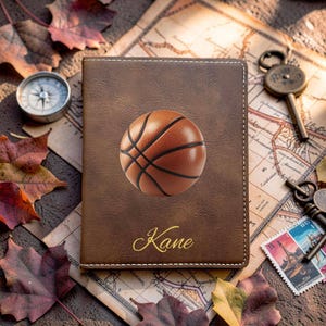 May include: A brown rectangular passport holder featuring a centered basketball graphic and the name Kane printed in gold script. The cover has visible white stitching along the edges and a smooth, leather-like texture with a matte finish.