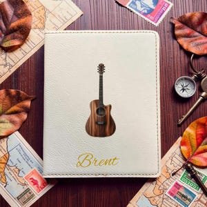 May include: A cream-colored passport holder with a textured surface featuring a centered illustration of a brown acoustic guitar and the name Brent printed in gold cursive script at the bottom center of the rectangular cover.