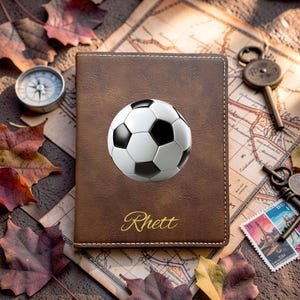 May include: A brown leather passport holder featuring a white and black soccer ball graphic centered on the front cover. The name Rhett is embossed in gold cursive script below the soccer ball. The cover has visible white stitching along the edges.