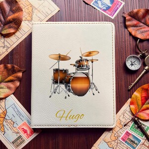 May include: A cream-colored rectangular passport holder featuring a printed graphic of a brown and black drum set in the center. The name Hugo is printed in a gold cursive script font below the drum set illustration on the textured cover.