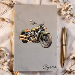 Peut inclure: Un carnet gris clair à texture marbrée orné d'un graphique de moto verte et dorée au centre, avec le nom Cyrus imprimé en écriture noire en bas, accompagné d'un stylo en métal doré posé sur le côté droit.