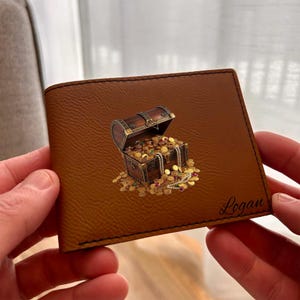 May include: A brown bi-fold wallet featuring a printed graphic of an open wooden treasure chest overflowing with gold coins, jewels, and a pearl necklace. The name Logan is printed in a black cursive script in the bottom right corner.