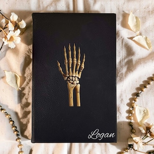 May include: A rectangular black notebook with a textured cover featuring a centred illustration of a human skeletal hand in beige and brown tones. The name Logan is printed in a white cursive script in the bottom right corner of the cover.