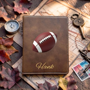 May include: A brown rectangular passport holder featuring a centered graphic of a brown and white American football and the name Hank printed in gold cursive script. The cover has visible white stitching along the edges of the brown material.