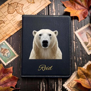 Könnte beinhalten: Ein dunkelblauer Reisepasshalter mit einem zentrierten, realistischen Porträt eines weißen Eisbären. Unter dem Bären ist der Name Reid in einer goldenen Schreibschrift aufgedruckt. Der Einband hat sichtbare weiße Nähte an den Rändern.