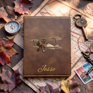 May include: A brown rectangular passport holder featuring a printed image of a vintage biplane in the center and the name Jesse written in gold cursive script below the aircraft. The cover has visible white stitching along the edges.