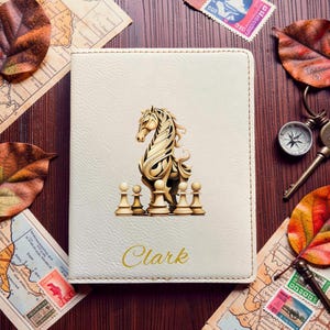 May include: A cream-colored passport holder featuring a central graphic of a stylized horse surrounded by several chess pawns. The name Clark is printed in gold cursive script at the bottom of the cover, which has visible stitching along the edges.