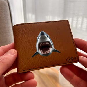 Peut inclure: Un portefeuille bifold marron clair avec une illustration centrale d'un requin la gueule ouverte, montrant des dents acérées. Le prénom Neil est inscrit en écriture cursive noire dans le coin inférieur droit du portefeuille aux coutures visibles.