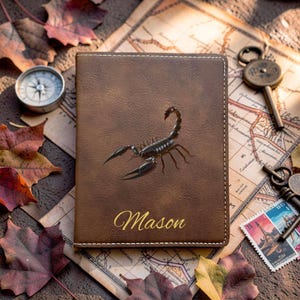 Pode incluir: Um porta-passaporte retangular castanho com um gráfico central de um escorpião preto e o nome Mason impresso em letra cursiva dourada. A capa apresenta costuras brancas visíveis ao longo das bordas e uma superfície texturizada.