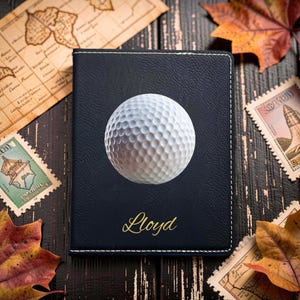 May include: A dark navy blue passport holder featuring a white dimpled golf ball graphic centered on the front cover. The name Lloyd is printed in gold cursive script below the golf ball. The cover has visible white stitching along the edges.