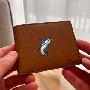 Peut inclure: Un portefeuille pliable marron clair à la surface texturée orné d'une illustration graphique d'un marlin bleu et argent au centre et du nom Dean imprimé en écriture cursive noire dans le coin inférieur droit avec des coutures noires.