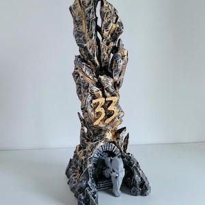 May include: A tall, vertical sculpture with a jagged, rock-like texture finished in black and gold paint. The number 33 is prominently displayed in gold on the front. At the base, there is a small arched entryway containing a light grey figure.