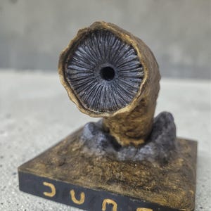May include: A detailed sculpture of a sandworm from the Dune series. The worm is a tan colour with a dark grey interior and sits on a black base with the word "DUNE" in gold lettering.
