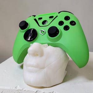Op de afbeelding: Een felgroene gamecontroller met zwarte knoppen en accenten rust op een witte buste. Op de controller staan de woorden "BACK" en "START". De buste is wit en heeft een gedetailleerd gezicht.
