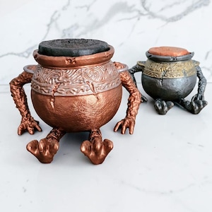 Puede incluir: Dos figuras decorativas con forma de olla con brazos y piernas. La olla más grande es de color cobre con una tapa oscura, mientras que la más pequeña es plateada y dorada con una tapa naranja. Ambas tienen texturas detalladas.