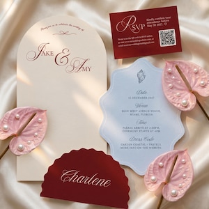 May include: A wedding stationery set featuring an arched cream invitation with the text Jake & Amy, a light blue scalloped card with event details, a dark red RSVP card with a QR code, a dark red name card, and three pink anthurium flowers.