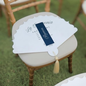 Pode incluir: Um programa de casamento branco em forma de leque com bordas onduladas e uma faixa azul marinho "Reserved". O programa está em uma cadeira com uma almofada bege e uma estrutura de madeira. Uma borla dourada pende da parte inferior.