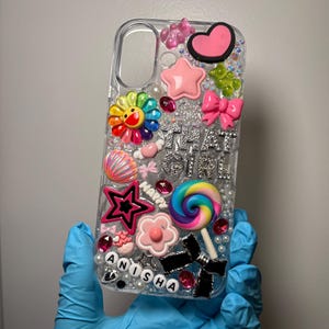 May include: Clear phone case decorated with various charms, including a pink heart, star, bow, and a rainbow lollipop. The case has the text "THAT GIRL" and the name "ANISHA" spelled out in beads. The case is held by a person wearing blue gloves.