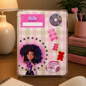 Pode incluir: Uma capa transparente para tablet com um padrão xadrez rosa e verde. A capa apresenta um gráfico de boneca, autocolantes e o texto "Hello my name is". Elementos decorativos: ursinhos de goma, um CD e um rádio rosa.