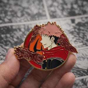 May include: A circular enamel pin featuring an anime character. The pin has a gold edge and shows a character with pink hair, a red cloak, and a black uniform. The background is red and orange.