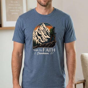 May include: A heather blue short-sleeve t-shirt featuring a graphic of a hiker on a mountain peak against a sunset. Below the graphic, the text reads "Walk by FAITH" followed by "2 Corinthians 5:7" in a combination of bold and script fonts.