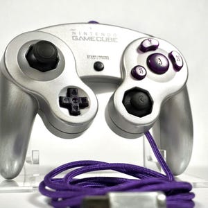 Puede incluir: Mando plateado de Nintendo GameCube con botones morados y cable morado. El mando tiene un joystick negro y una cruceta. El texto "NINTENDO GAMECUBE" está impreso en la parte superior.