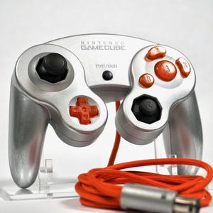 May include: Silver Nintendo GameCube controller with orange buttons and a black joystick. The controller has a red cross-shaped directional pad and a coiled orange cable with a silver connector. The GameCube logo is visible on the top.