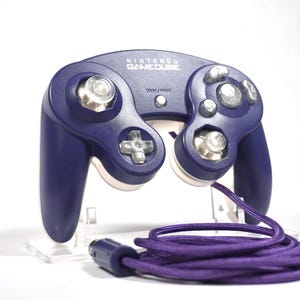May include: A purple Nintendo GameCube controller with a matching purple cord. The controller has a classic design with multiple buttons and analog sticks. The GameCube logo is visible on the top.