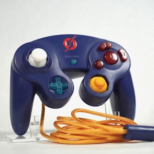 May include: A dark blue video game controller featuring a white analog stick, a yellow C-stick, a teal D-pad, and red action buttons. The center displays a red lightning bolt logo above the text "THRA/PARA" and an orange braided cable.