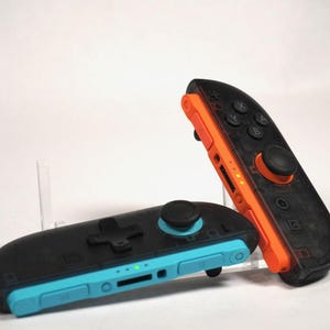 May include: Two video game controllers featuring a translucent black shell. One controller has a bright orange side rail and buttons, while the other features a vibrant cyan blue side rail and buttons. Both units include a black analog joystick.