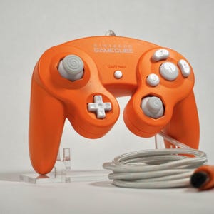 May include: Orange Nintendo GameCube controller with gray buttons and a white cross-shaped directional pad. The controller has the text "NINTENDO GAMECUBE" and "START/PAUSE" printed on it. A gray and white cable is coiled below the controller.