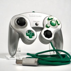 Puede incluir: Mando plateado de Nintendo GameCube con botones verdes y negros y un cable verde. El mando tiene un diseño clásico con una cruceta, palancas analógicas y varios botones. El logotipo de GameCube es visible en la parte superior.